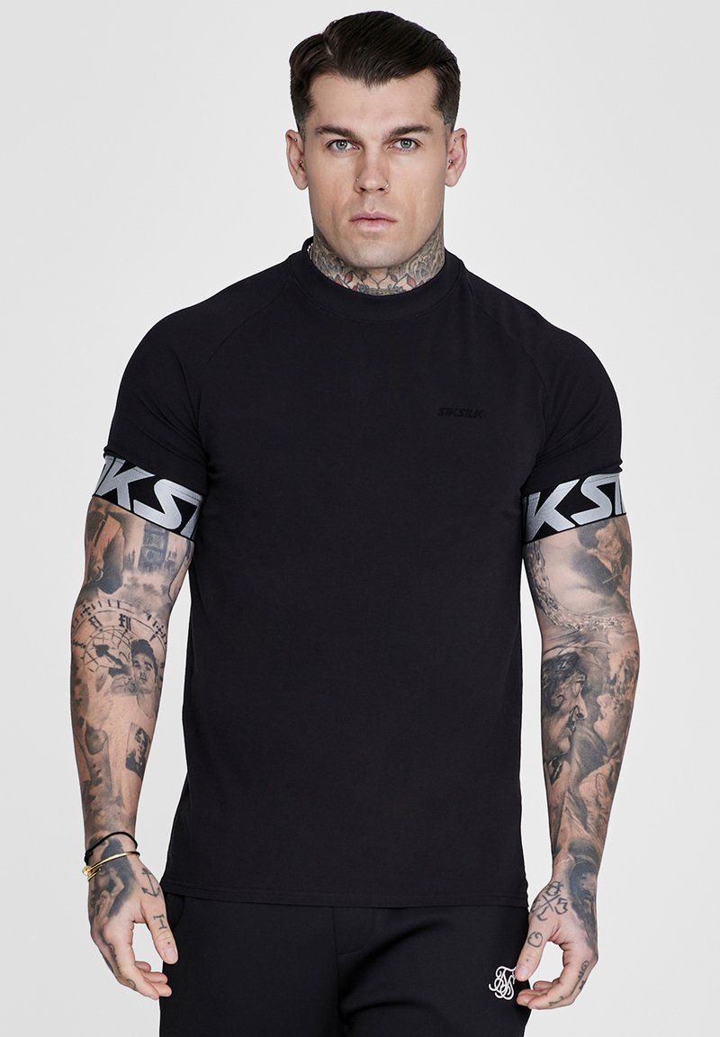 Homme tatoué portant un T-shirt noir ajusté et un short noir, debout avec une expression neutre sur un fond uni.