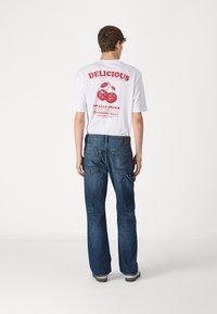Camiseta blanca con texto rojo y gráficos de cerezas en la parte trasera, combinada con jeans de campana azules. Los jeans tienen una tela texturizada y bolsillos.