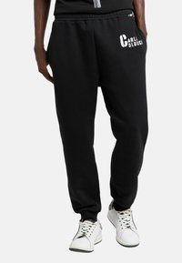 Mand, der bærer sorte joggerbukser med "Carlo Colucci" tekst på venstre lår og hvide sneakers på en hvid baggrund.