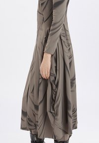 Vestido largo y ajustado en un tejido taupe suave, con detalles drapeados y un patrón texturizado, de mangas largas y una silueta fluida.