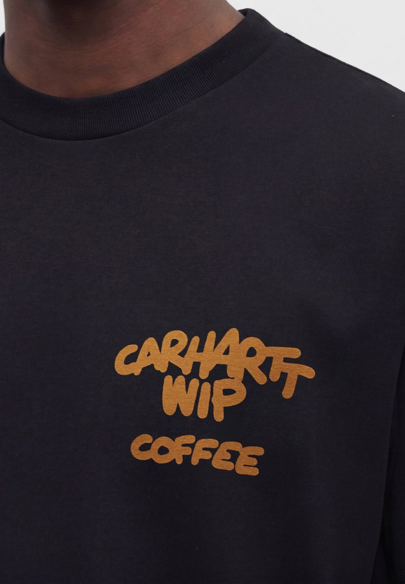Schwarzes Crewneck-Shirt mit senfgelbem Text "CARHARTT WIP COFFEE" auf der Brust, getragen von einer Person mit dunkler Haut.