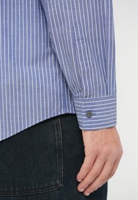 Δεξί χέρι ατόμου που φοράει μπλε και λευκή ριγέ πουκαμίσα με μακριά μανίκια, κουμπωμένο στο μανίκι, resting δίπλα από την τσέπη των σκούρων τζην.