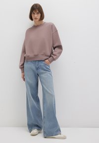 Light mauve oversized sweatshirt with ribbed hem, paired with high-waisted, wide-leg denim jeans in a faded blue wash. Casual footwear.