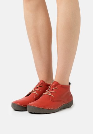 Underben iført røde læderankelstøvler med beige snørebånd og mørkebrune gummisåler, stående på en hvid baggrund.