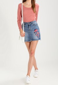 Jupe en denim mini avec ourlet effiloché et broderie florale colorée sur le devant, associée à un top rose à manches longues et des baskets blanches.