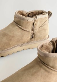 Botas de tobillo de ante beige con cremallera en el lateral, forro de suave pelaje y suela de goma texturizada con un patrón de tacos.