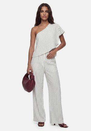 Vrouw draagt een witte top met één schouder en een broek met zwarte verticale strepen, houdt een bordeaux gevlochten handtas en bijpassende sandalen met hak vast.