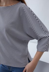 Maglietta a righe bianca e nera, con scollatura a barca, vestibilità ampia e maniche a tre quarti con fessure laterali. Tessuto morbido e testurizzato.