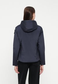 Marineblauwe waterdichte jas met een capuchon, lange mouwen en een gladde, lichte textuur. Heeft een schouderpatch voor detail. Achteraanzicht weergegeven.