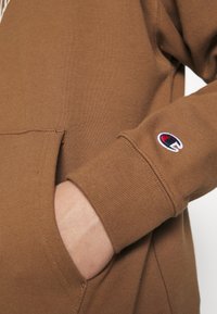 Sudadera marrón con bolsillo tipo canguro y puños acanalados. Incluye un pequeño logo bordado en la manga. Material de algodón.