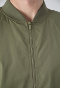 Giacca bomber verde oliva con colletto a coste, zip a lunghezza intera e trama del tessuto liscia. Nessun motivo o dettaglio aggiuntivo visibile.