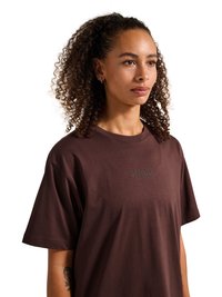 Vrouw met krullend haar die een donkerbruine T-shirt draagt met "HALO" op de borst gedrukt, staand tegen een witte achtergrond.