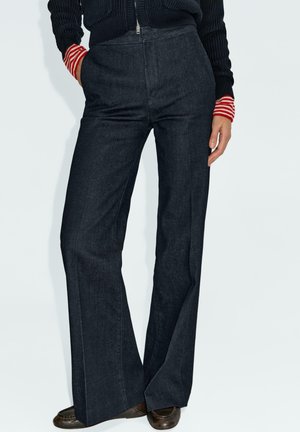 Mörka denimflared byxor med hög midja, med en slät textur och subtila sömmar. Sammanfogas med en svart stickad tröja och rödrandiga manschetter.