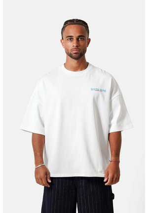 Homme avec des cheveux tressés, portant un t-shirt blanc oversize avec un petit texte sur la poitrine et un pantalon sombre à fines rayures, debout devant un fond blanc.