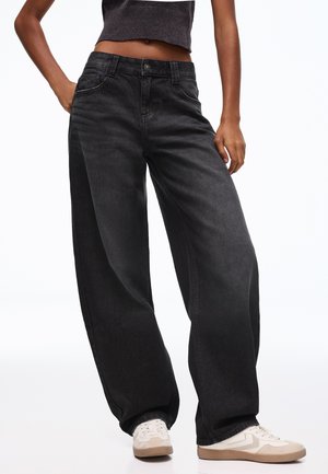 Persona che indossa jeans neri larghi, top corto scuro e scarpe da ginnastica bianche, in piedi con una mano in tasca su sfondo bianco.