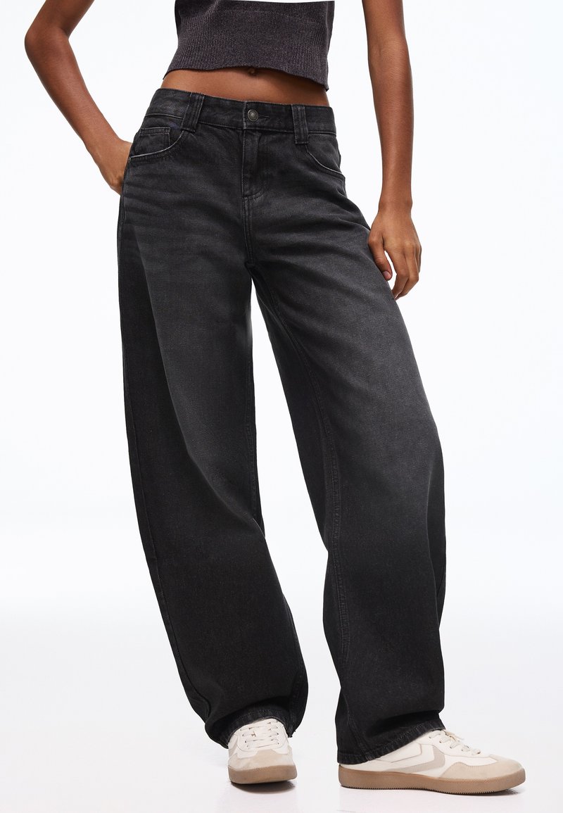 Person, die lockere schwarze Jeans, ein kurzes dunkles Oberteil und weiße Turnschuhe trägt, steht mit einer Hand in der Tasche vor weißem Hintergrund.