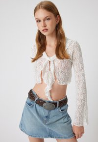 Valkoinen pitsinen crop-top, jossa on pitkät hihat, solmimisetu ja röyhelöhelma. Se on yhdistetty vaalean denim-minihelmaan ja punottuun ruskeaan vyöhön.