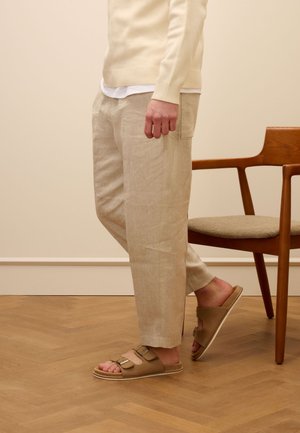 Pantaloni di lino beige con una vestibilità comoda e tasca laterale, abbinati a sandali marroni con cinturino a fibbia. Sedia in legno sullo sfondo.