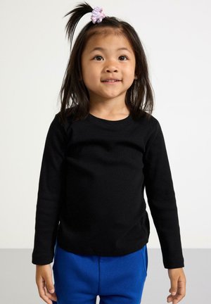 Svart ribbad långärmad skjorta i bomull, tillsammans med blå byxor. Barnet har en hästsvans med en ljuslila håraccessoar.