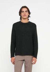 Zwarte gebreide trui met een ronde hals, geribde manchetten en zoom; heeft een gestructureerd patroon over de gehele trui. Draagt een lichtbruine broek.