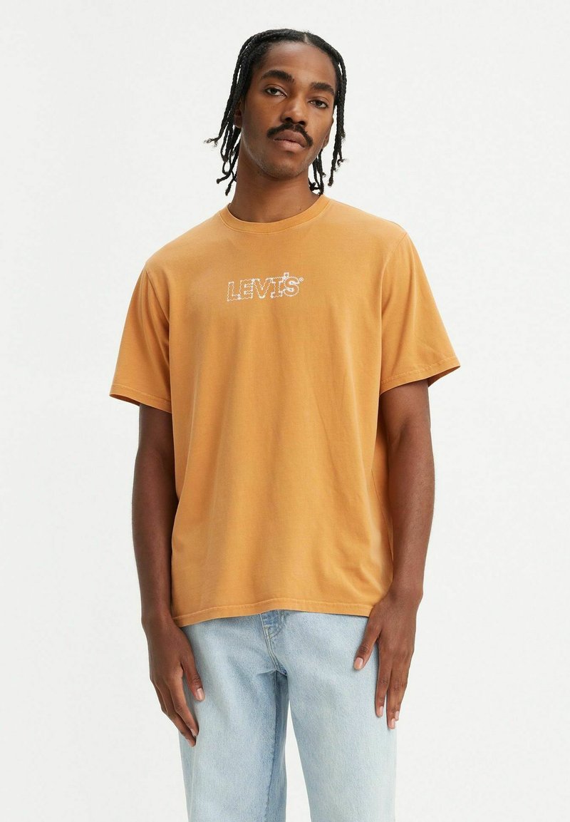 Camiseta de manga corta de color mostaza, hecha de algodón, con un gráfico blanco del logo "LEVI'S" en el centro, combinada con jeans azul claro.