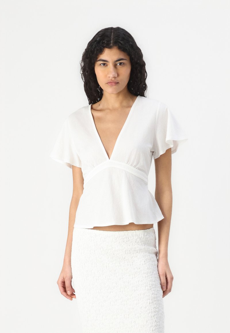 Hvit kortermet topp med dyp V-hals, en tettsittende midje og en peplum-bunn. Laget av lett stoff med en glatt tekstur.