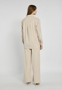 Beżowa koszula z korduroju i szerokie spodnie o luźnym kroju. Posiada mankiety na guziki i fałdę z tyłu dla dodatkowych detali.