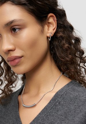 Frau mit lockigem Haar, die silberne Creolen, eine silberne Halskette und ein graues V-Ausschnitt-Oberteil vor schlichtem Hintergrund trägt.