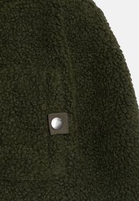 Grüner Fleece-Stoff mit strukturierter Oberfläche, versehen mit einem dunklen Stoffetikett und einem silbernen Knopfdetail. Weich, warm und visuell einheitlich.