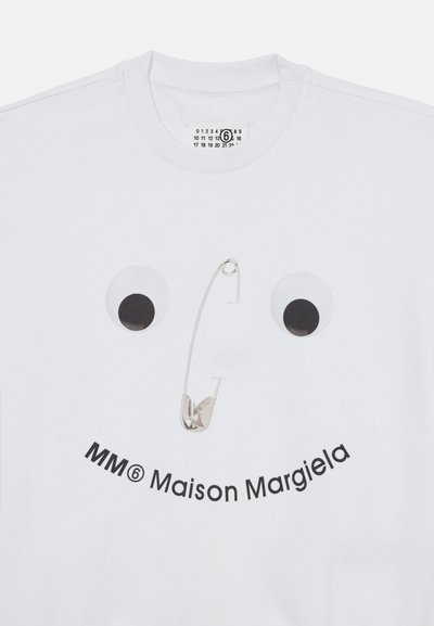 Balts džemperis ar izdrukātu sejas dizainu, kuram ir lieli googlējoši acis un drošības adata. Apakšā rakstīts "MM6 Maison Margiela" treknajiem melnajiem burtiem.