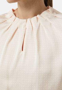 Helles pinkes Blusenshirt aus strukturiertem Stoff, mit gefälteltem Rundhalsausschnitt und kleinen Schlüsselöffnungen. Verfügt über ein dezentes geometrisches Muster.