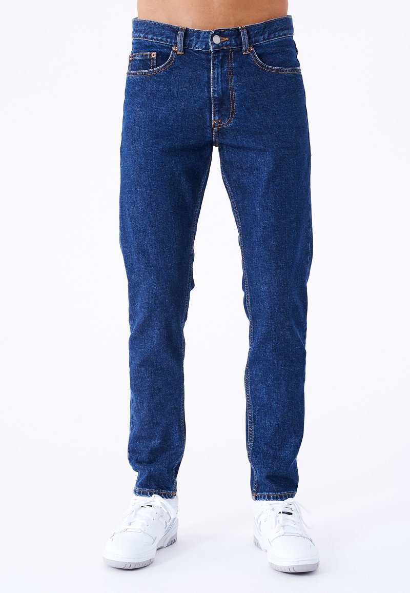 Jeans de mezclilla azul con corte recto, con cierre de botón, bolsillos estándar y costuras naranjas en contraste. Combinados con zapatillas blancas.