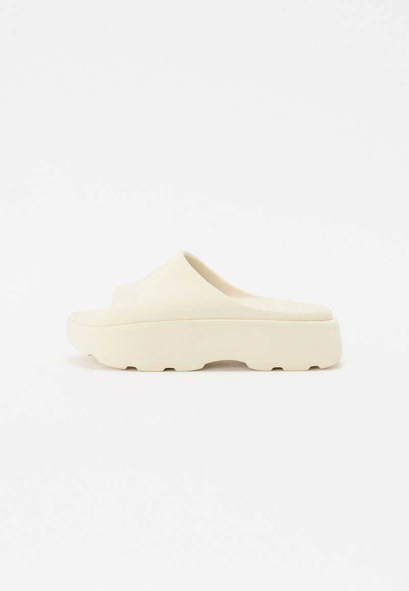 Beige slip-on sandaler med en slät gummiliknande yta, rund tå, tjock sula och strukturerad botten för grepp. Minimalistisk design.