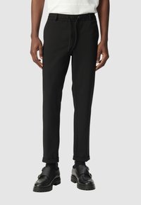 Uomo che indossa pantaloni slim-fit neri con risvolto e vita con cordino, abbinati a scarpe monk strap in pelle nera su sfondo grigio.