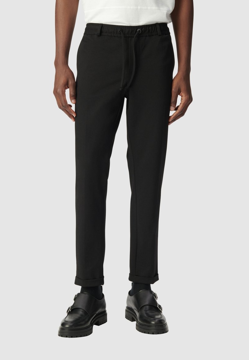 Uomo che indossa pantaloni slim-fit neri con risvolto e vita con cordino, abbinati a scarpe monk strap in pelle nera su sfondo grigio.