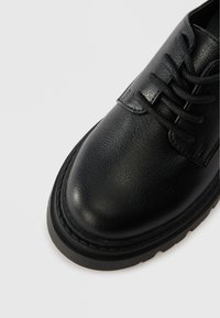 Pantof de piele neagră cu șireturi, cu un superior texturat, vârf rotund și o talpă groasă din cauciuc, având detalii de cusătură pe margine.