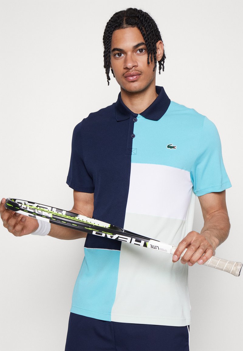 Polo shirt avec des blocs de couleur marine, turquoise et blanche, avec un col rabattu et le logo Lacoste. Un joueur tient une raquette de tennis.