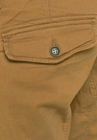 Tan fabric pant with a flap pocket, a metallic button with a four-hole design, and smooth texture, highlighting stitching details.