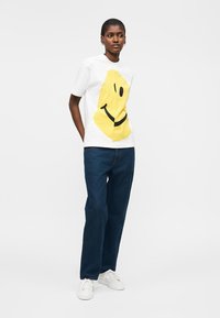 Femme debout les mains dans le dos, portant un T-shirt blanc avec un grand visage souriant jaune, un jean bleu foncé et des baskets blanches.
