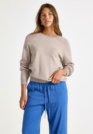 Beige strikkegenser med raglanermer, rund hals og ribbestrikkede mansjetter, sammen med løse blå bukser med snøring i livet.
