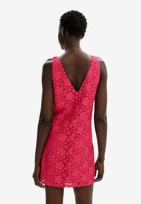 Modelka stojí zády k fotoaparátu, má na sobě živě růžové krajkové šaty s výstřihem do V, které předvádějí květinové vzory. Jednoduché pozadí.