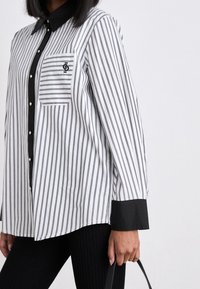 Chemise à rayures blanches et noires avec un col et des poignets noirs, dotée d'une poche poitrine et d'une fermeture à boutons. Texture de tissu lisse.