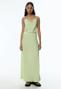 Cowl neck, sleeveless, floor-length dress in soft pastel green fabric. Features a fitted silhouette and smooth texture. Black platform sandals at feet.