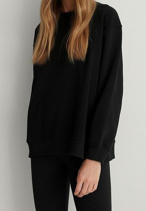 Person iført en løs sort langærmet sweatshirt og sorte bukser, med langt, lige lysbrunt hår, stående mod en ensfarvet væg.