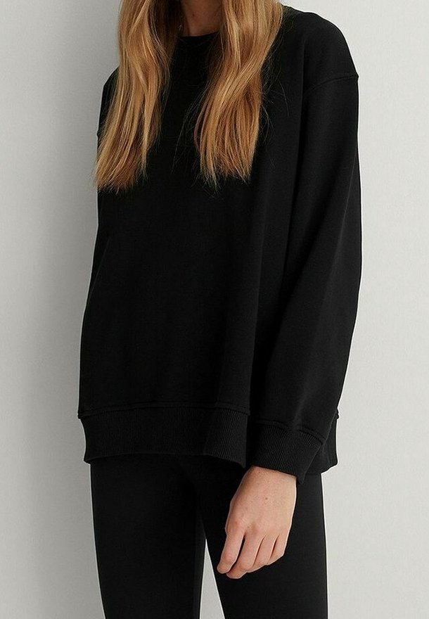 Personne portant un sweat-shirt noir ample à manches longues et un pantalon noir, avec de longs cheveux raides châtain clair, debout contre un mur uni.