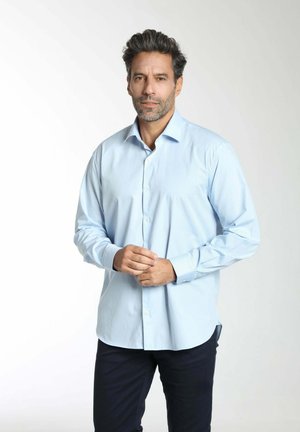 Homme aux cheveux foncés et à la barbe, portant une chemise bleue claire boutonnée et un pantalon foncé, debout les mains jointes devant un fond uni.