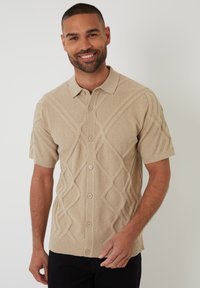 Polo de punto de manga corta beige con un patrón de diamantes texturizado, con cierre de botones y detalles de ribete en el dobladillo y el cuello.