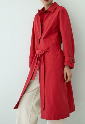 Donna che indossa un trench rosso lungo fino al ginocchio, legato in vita, abbinato a pantaloni beige a gamba larga su uno sfondo chiaro neutro.