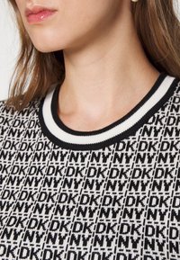 Gros plan d'une femme portant un pull à motifs noir et blanc avec le texte répété "DKNY" et un col rond rayé.