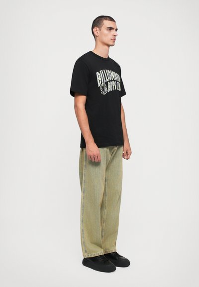Schwarzes T-Shirt mit weißem und grünem Text "Billionaire Boys Club", kombiniert mit hellgrünen, weit geschnittenen Jeans und schwarzen Sneakers.